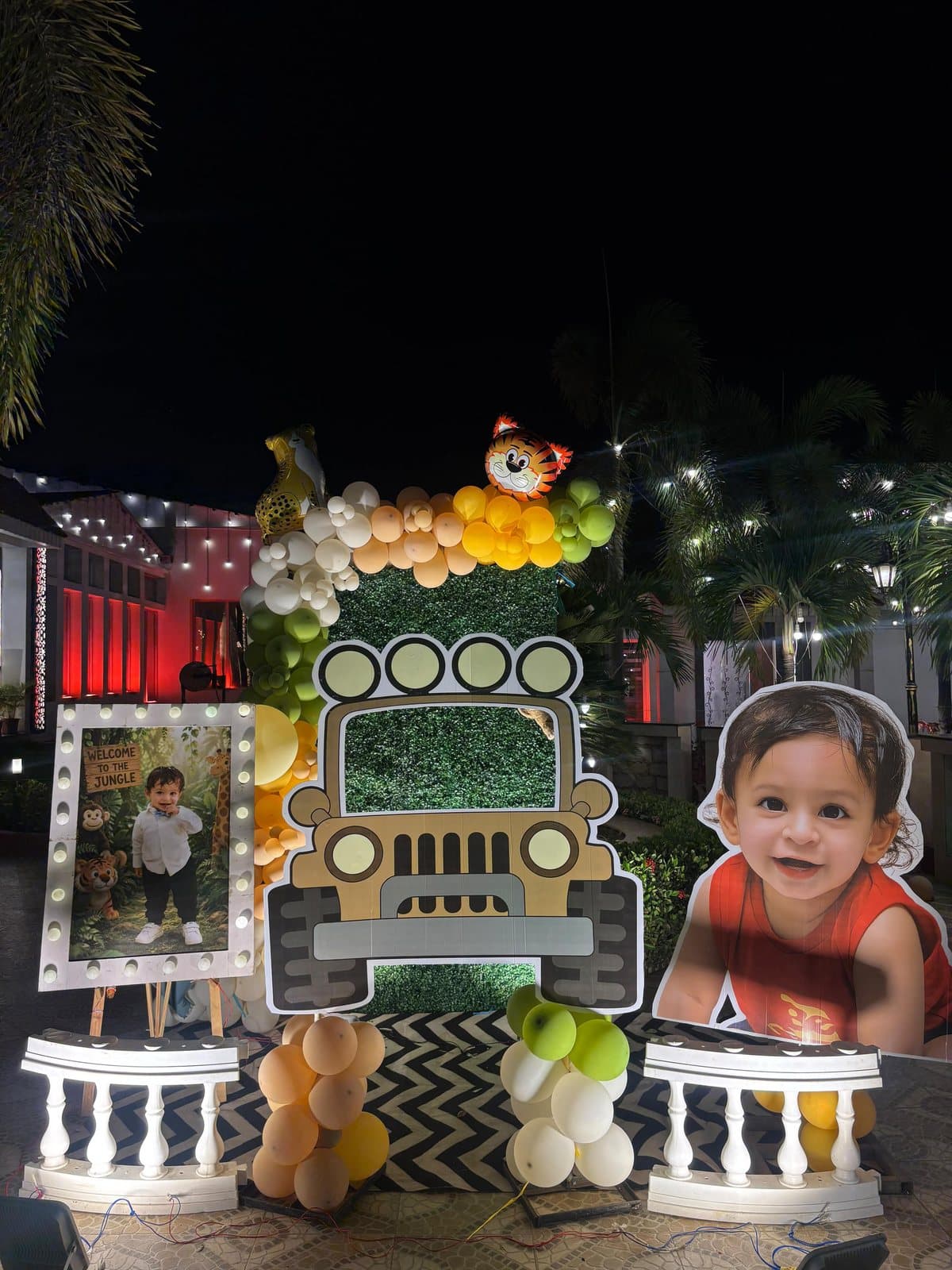 Night-time jungle safari birthday setup with a safari jeep cutout, green hedge wall and orange and green balloon garland