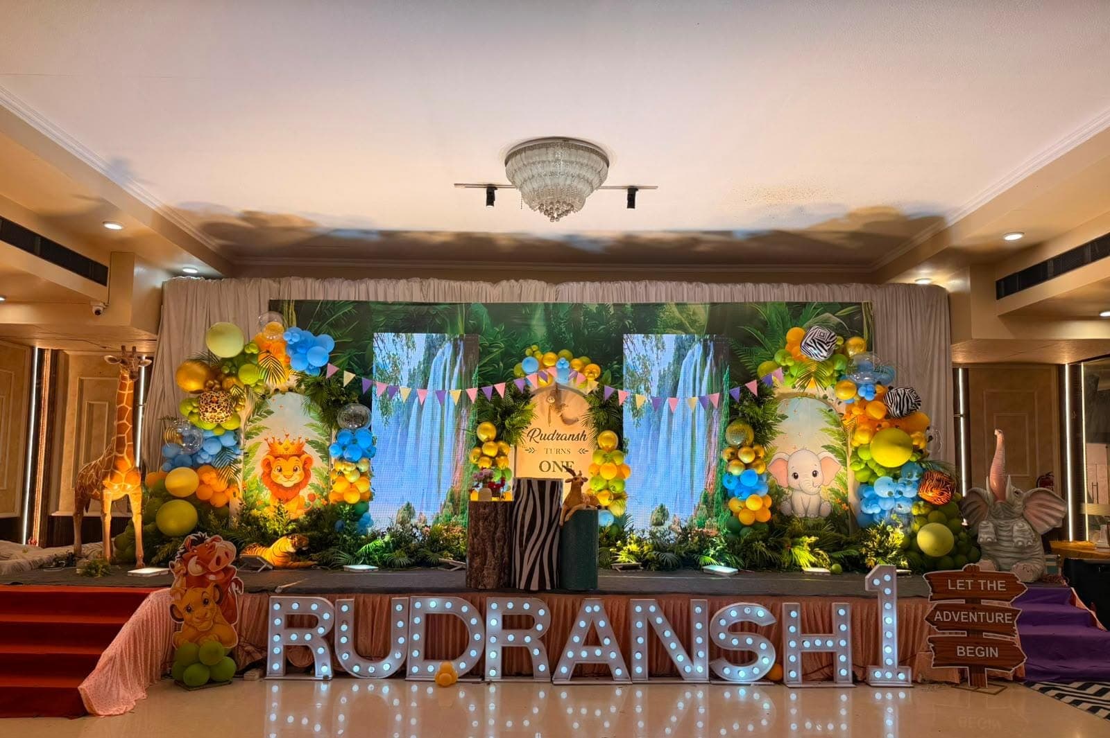 Rudransh's jungle-themed first birthday stage with illuminated marquee letters, balloon arches and safari animal props