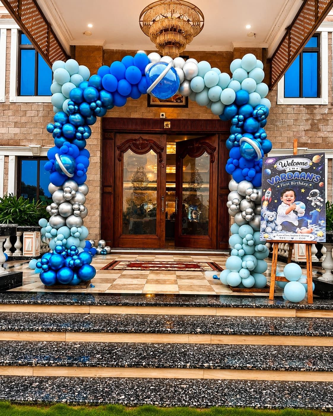Blue, sky-blue and silver balloon arch framing a villa entrance with a first birthday welcome board