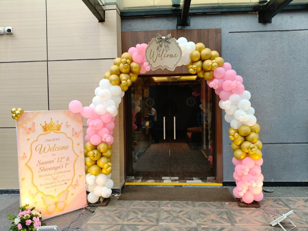 Pink, white and gold balloon entrance arch with a welcome board for a joint birthday celebration